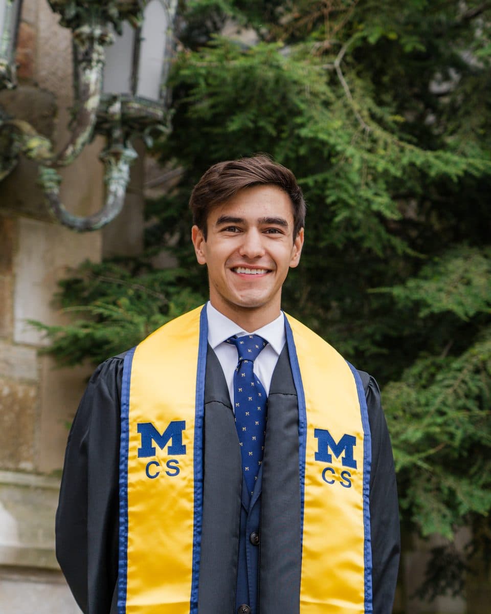 Neev smiling in a Michigan CS graduation stole outside a stone campus building